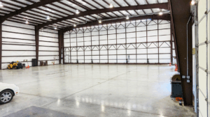 Empty large warehouse interior with high ceiling, concrete floor, and overhead lighting. Yellow machinery and traffic cones along wall.