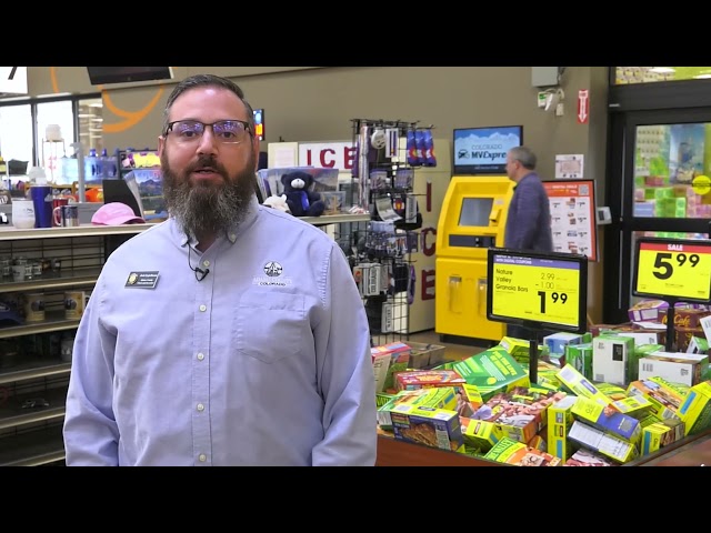 Man in a store with beard, wearing glasses and a uniform, standing near packed items and electronic signs displaying prices.