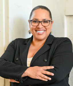 Dionne smiling confidently, arms crossed, wearing glasses and a black blazer.