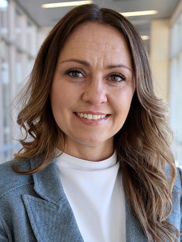 Nicci-Beauprez smiling in a bright hallway wearing a blue blazer and white shirt.