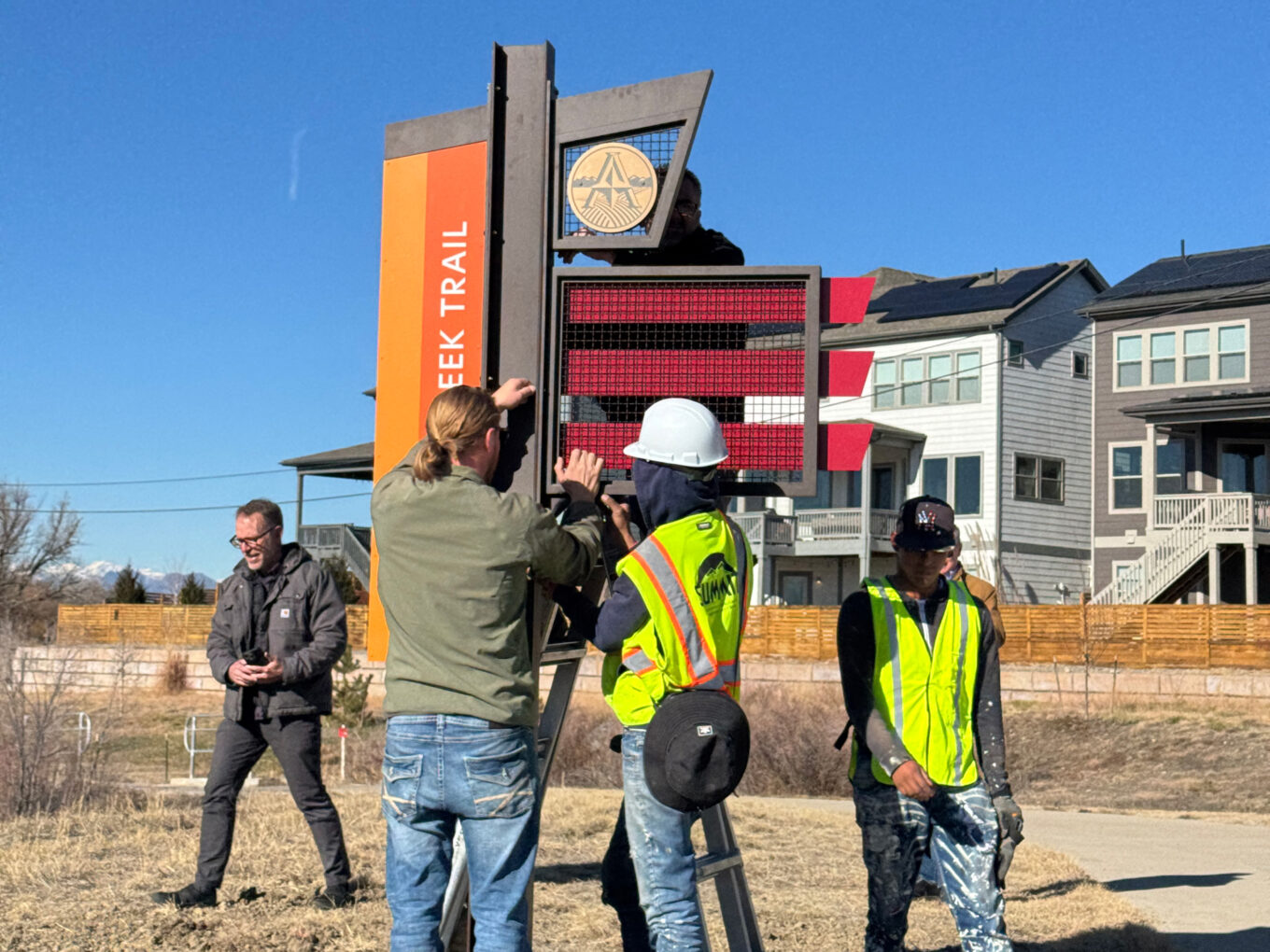 Workers installing Clear Creek Trail sign, wearing safety gear, blue sky background
