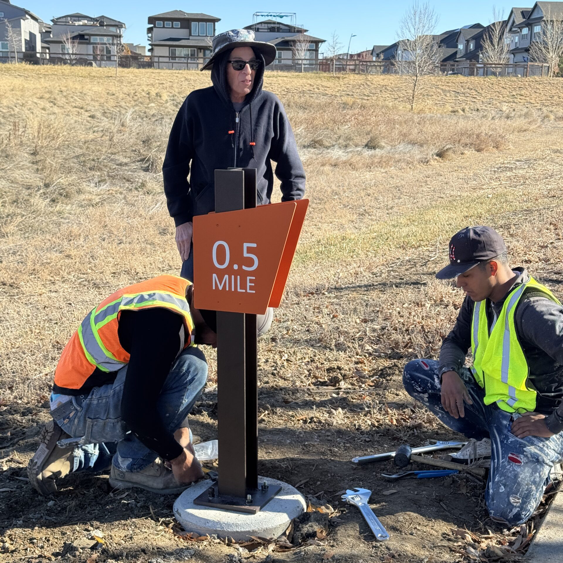 Three workers installing a "0.5 Mile" trail marker on a grassy path