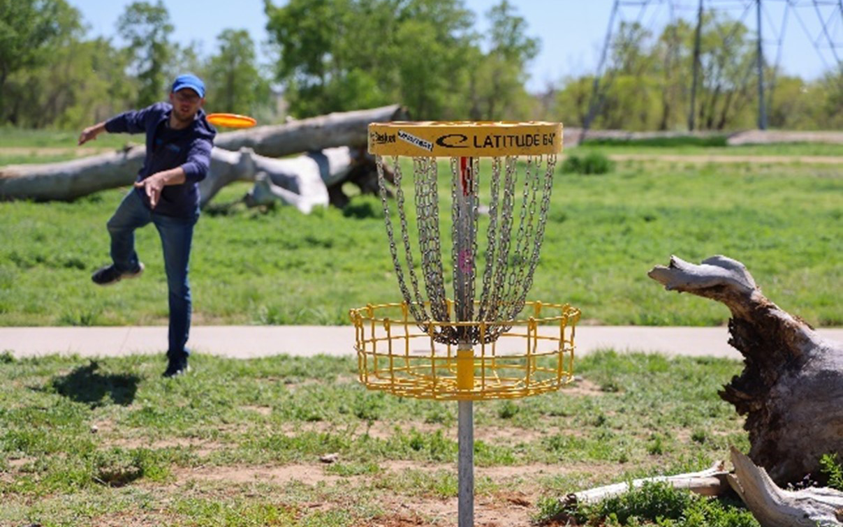 Person playfully throws a disc towards a disc golf basket in a grassy park setting