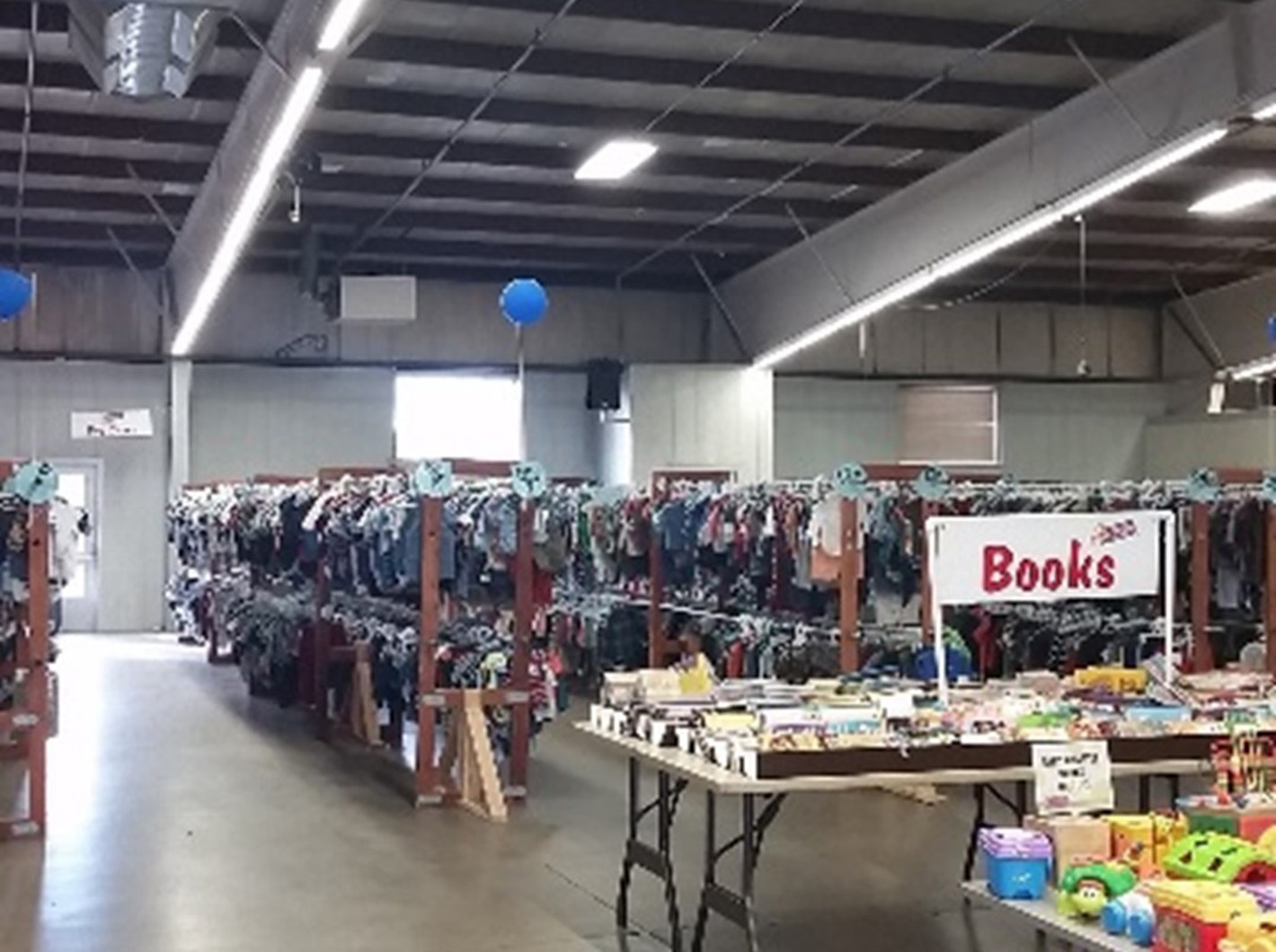 Rows of children's clothing racks and a table labeled "Books" in a warehouse sale setting.