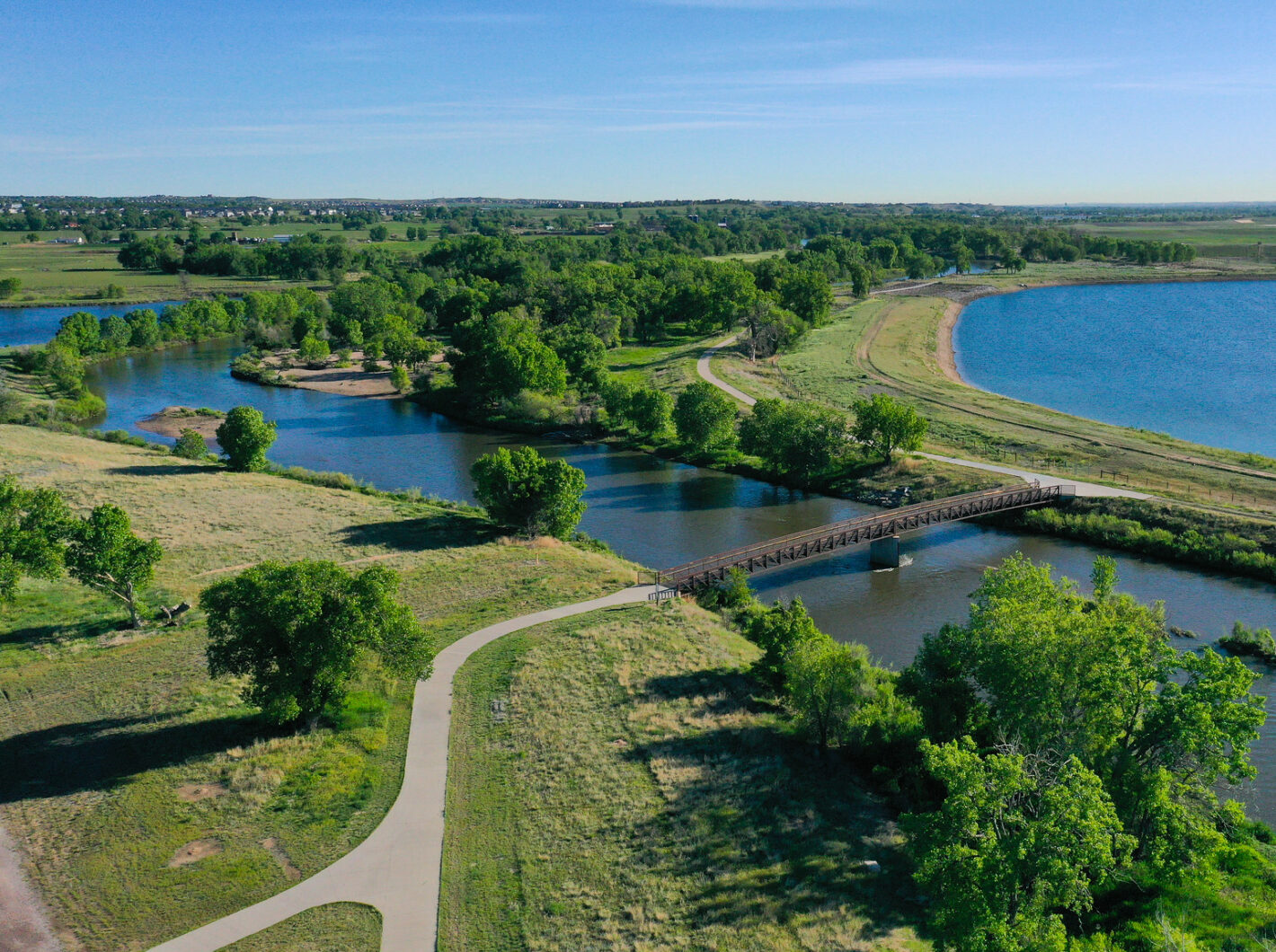 Aerial view of a scenic park with a winding path, bridge, trees, and a large lake.