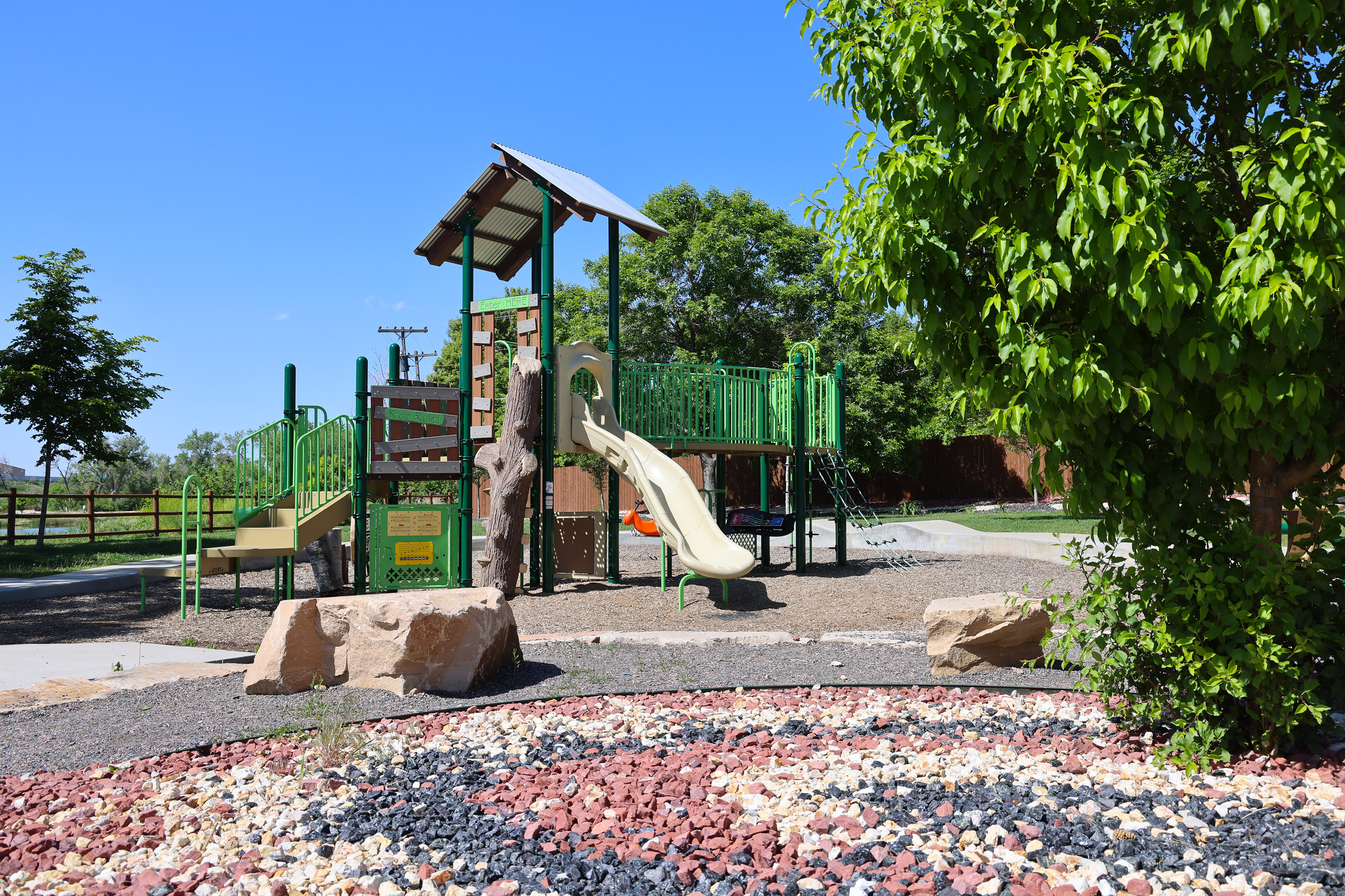 Playground with green structures, slide, climbing areas, surrounded by trees and rocks under a clear blue sky.