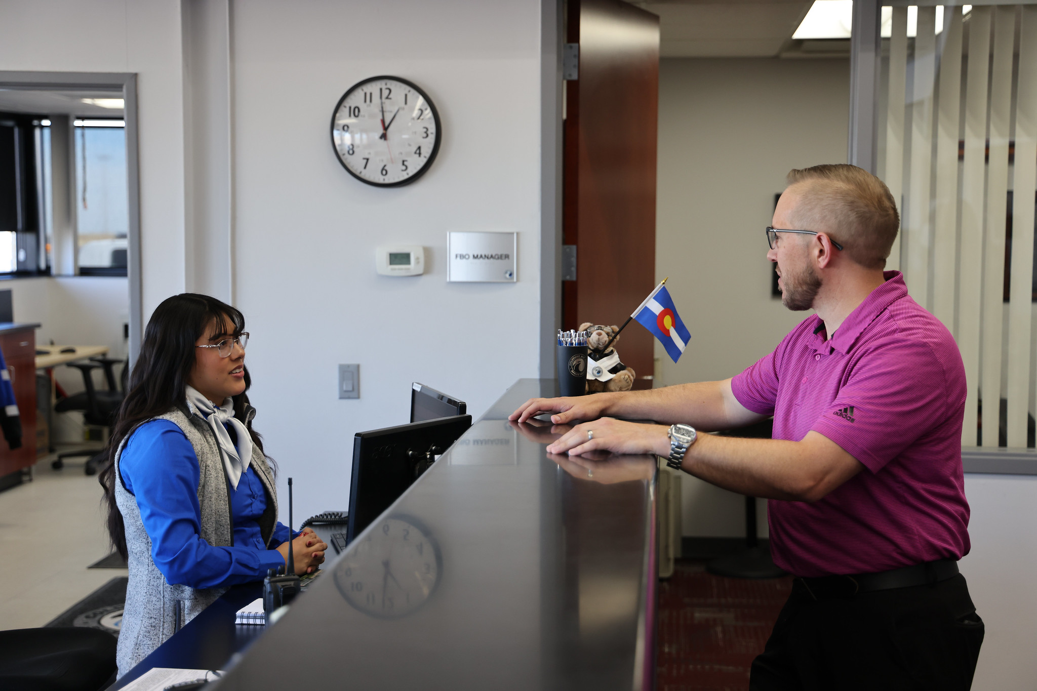 Man in pink shirt talking to receptionist by desk with clock labeled "FBO Manager" on wall.