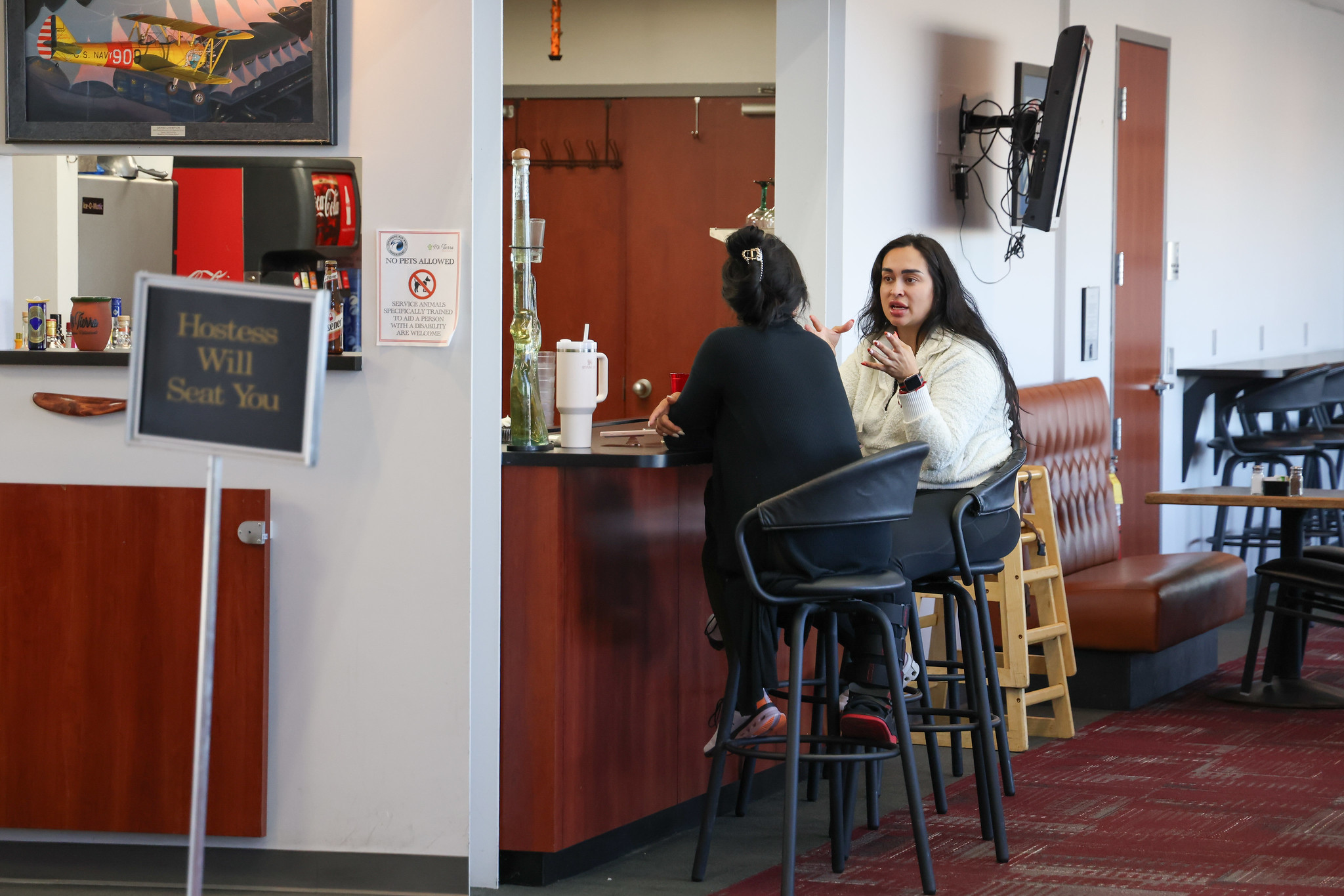 Two people seated at a bar, one gesturing while talking. Sign reads, "Hostess Will Seat You."
