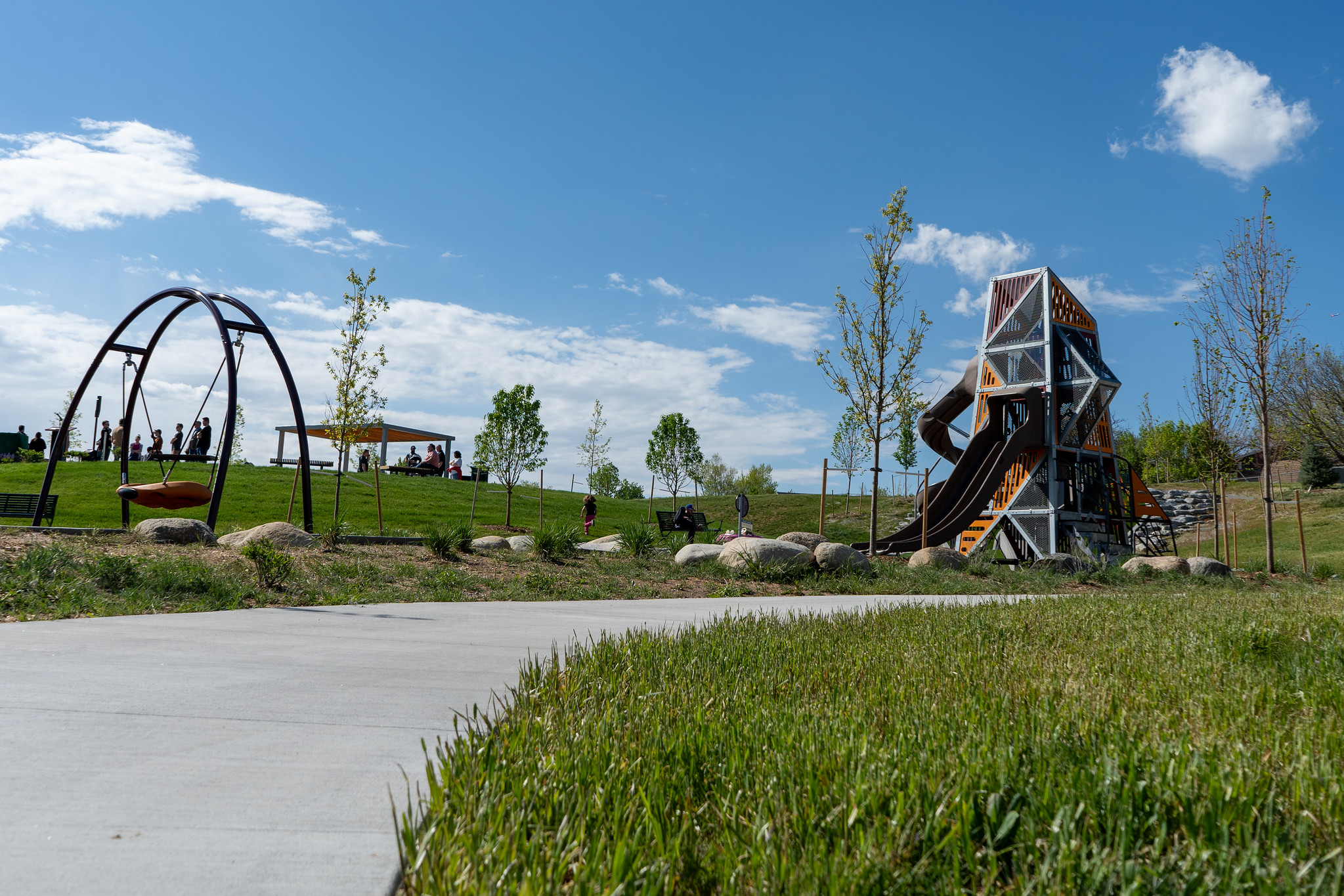 Sunny park scene with modern playground equipment, people sitting, and children playing.