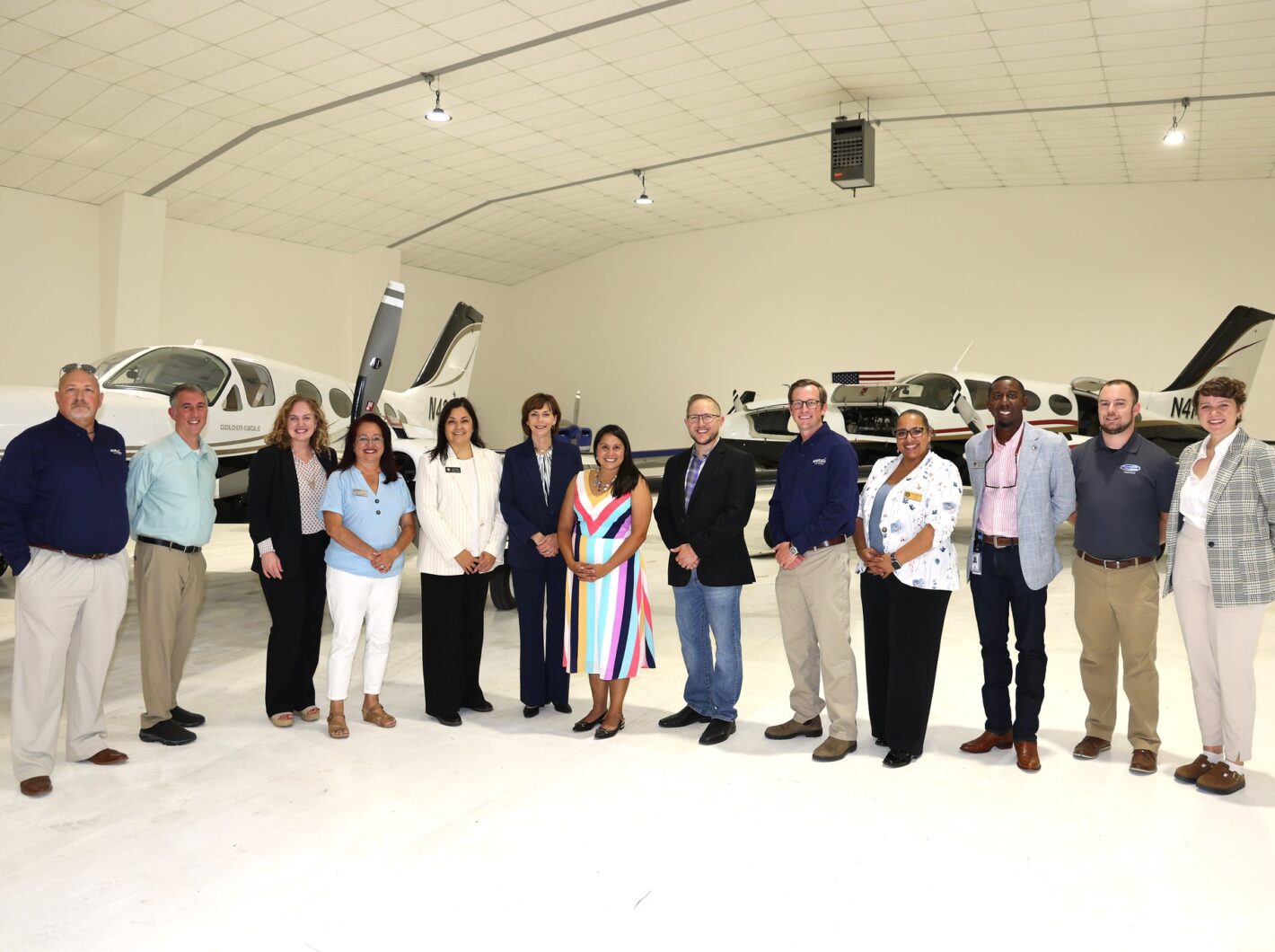 Group of thirteen people standing in front of two small airplanes inside a hangar, smiling and posing together