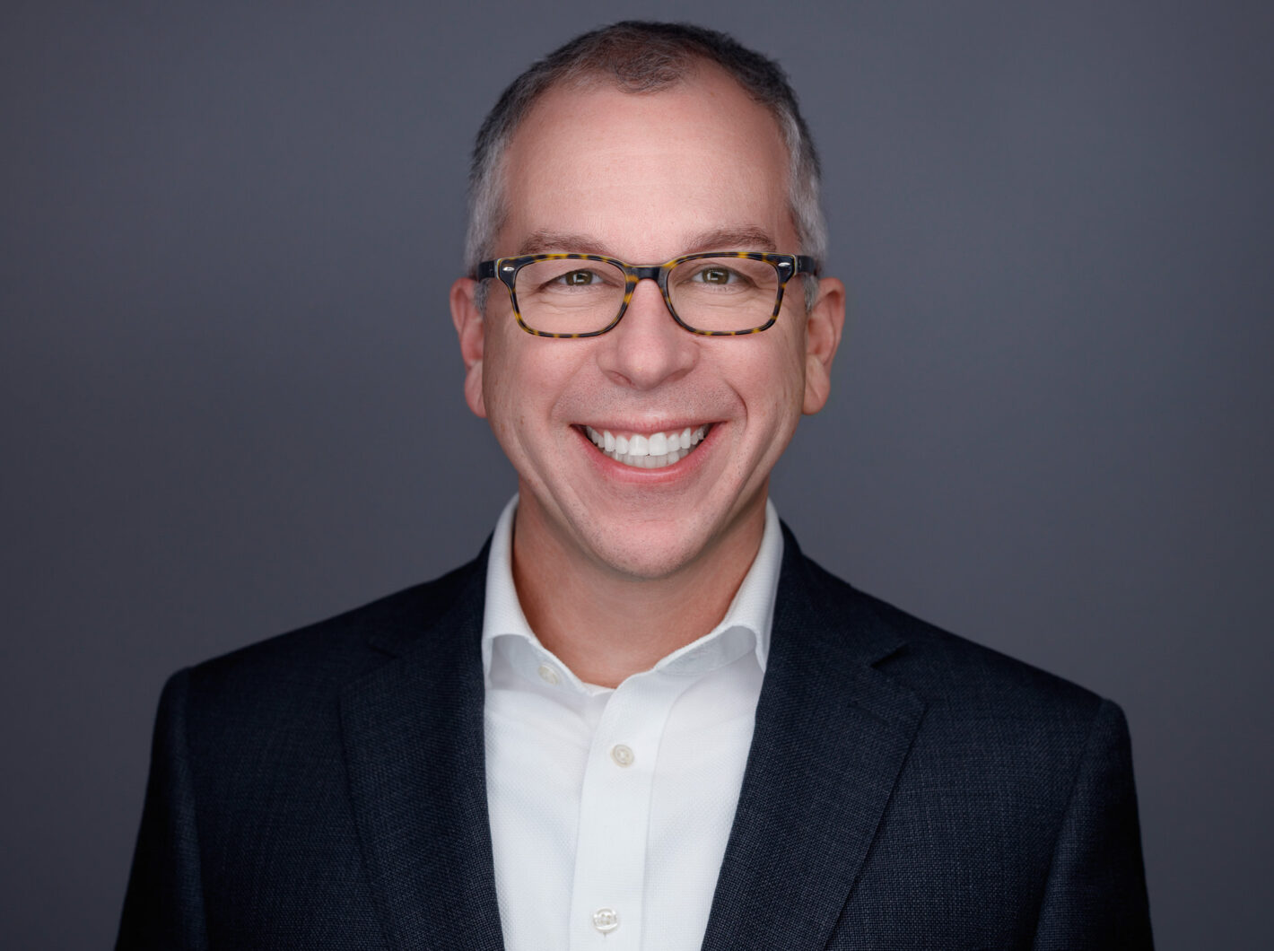 Jason-Schultz smiling, wearing glasses and a suit against a gray background