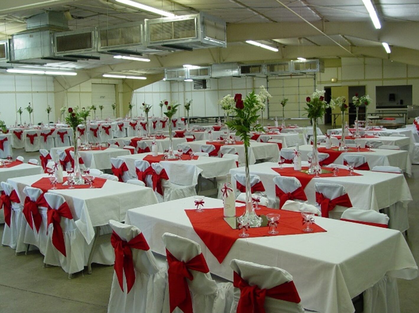 Event hall setup with tables draped in white cloths, red accents, and floral centerpieces.