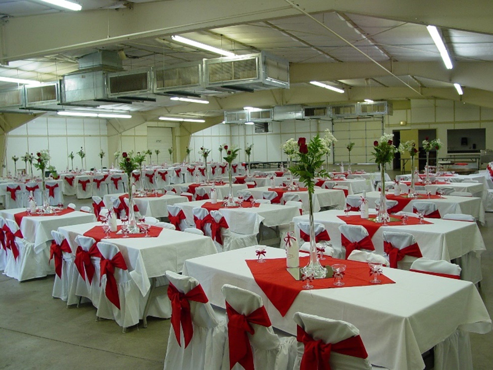 Event hall setup with tables draped in white cloths, red accents, and floral centerpieces.