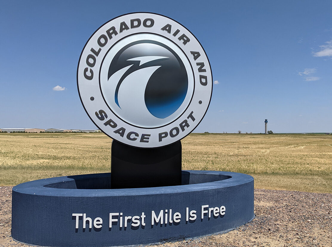 Colorado Air and Space Port sign with slogan "The First Mile Is Free" against clear sky and open field.