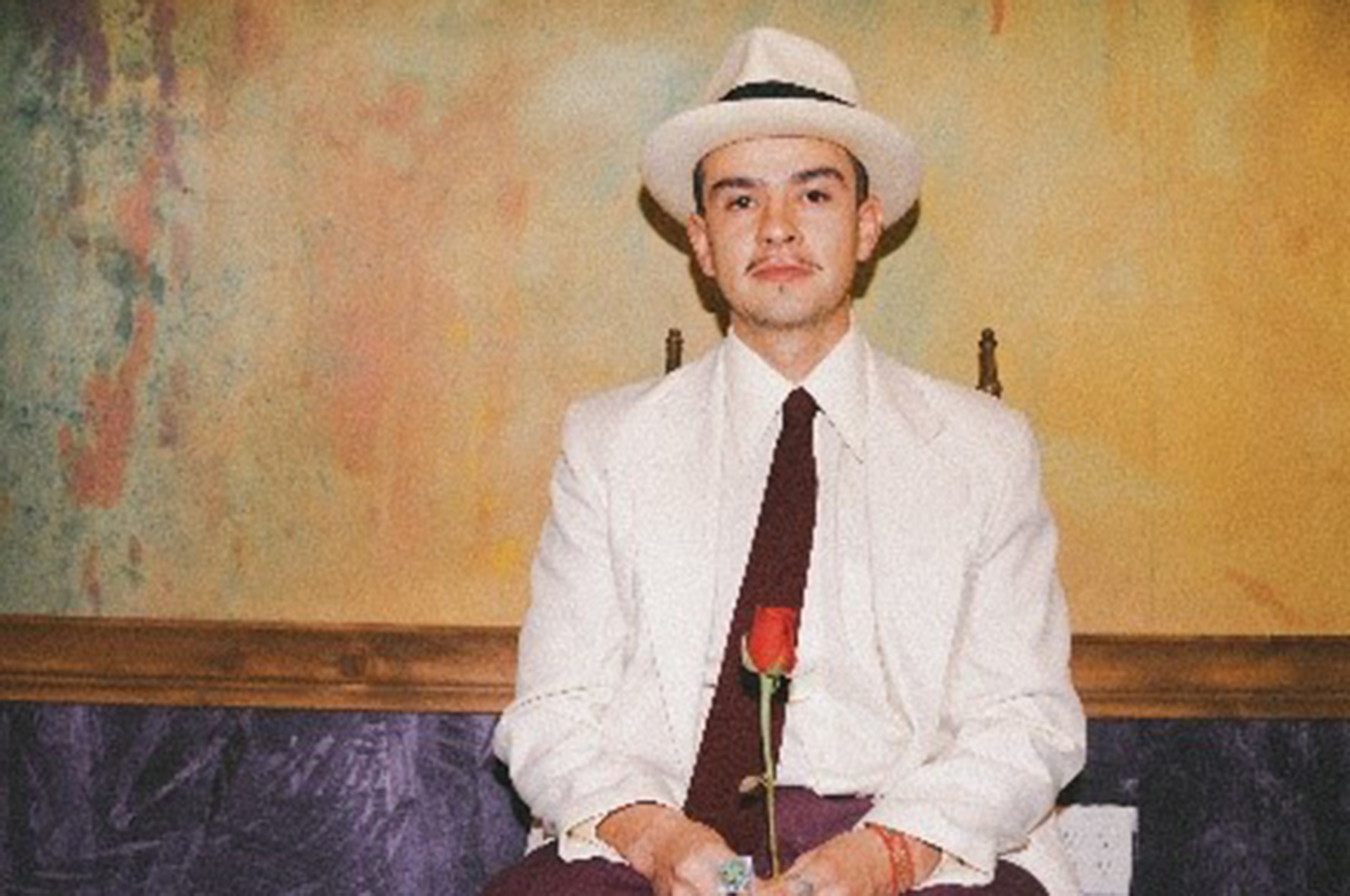DF.png: Person in a white suit and hat holding a rose, sitting against a colorful wall.