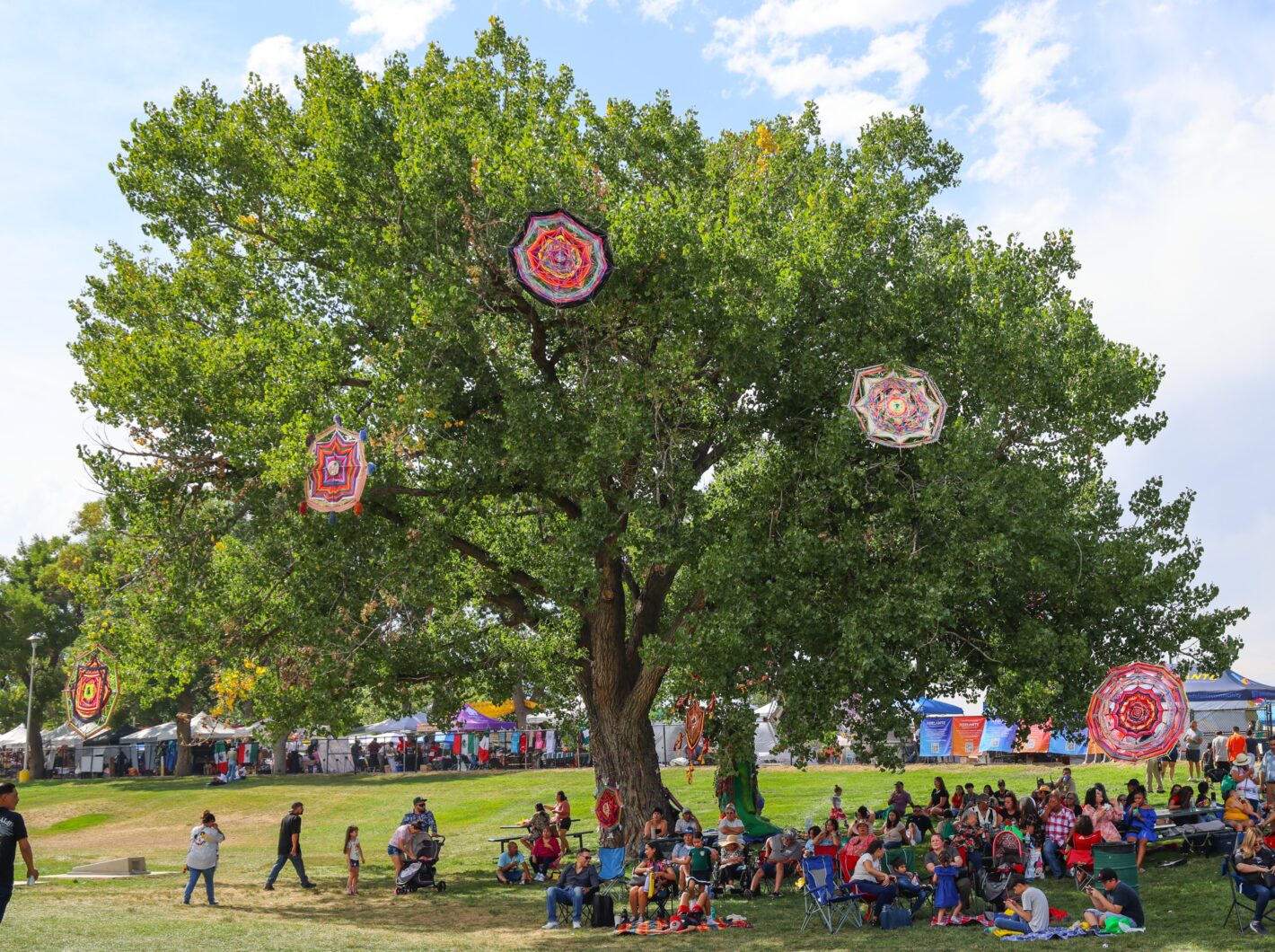 Large tree adorned with colorful patterns, people seated and walking in park.