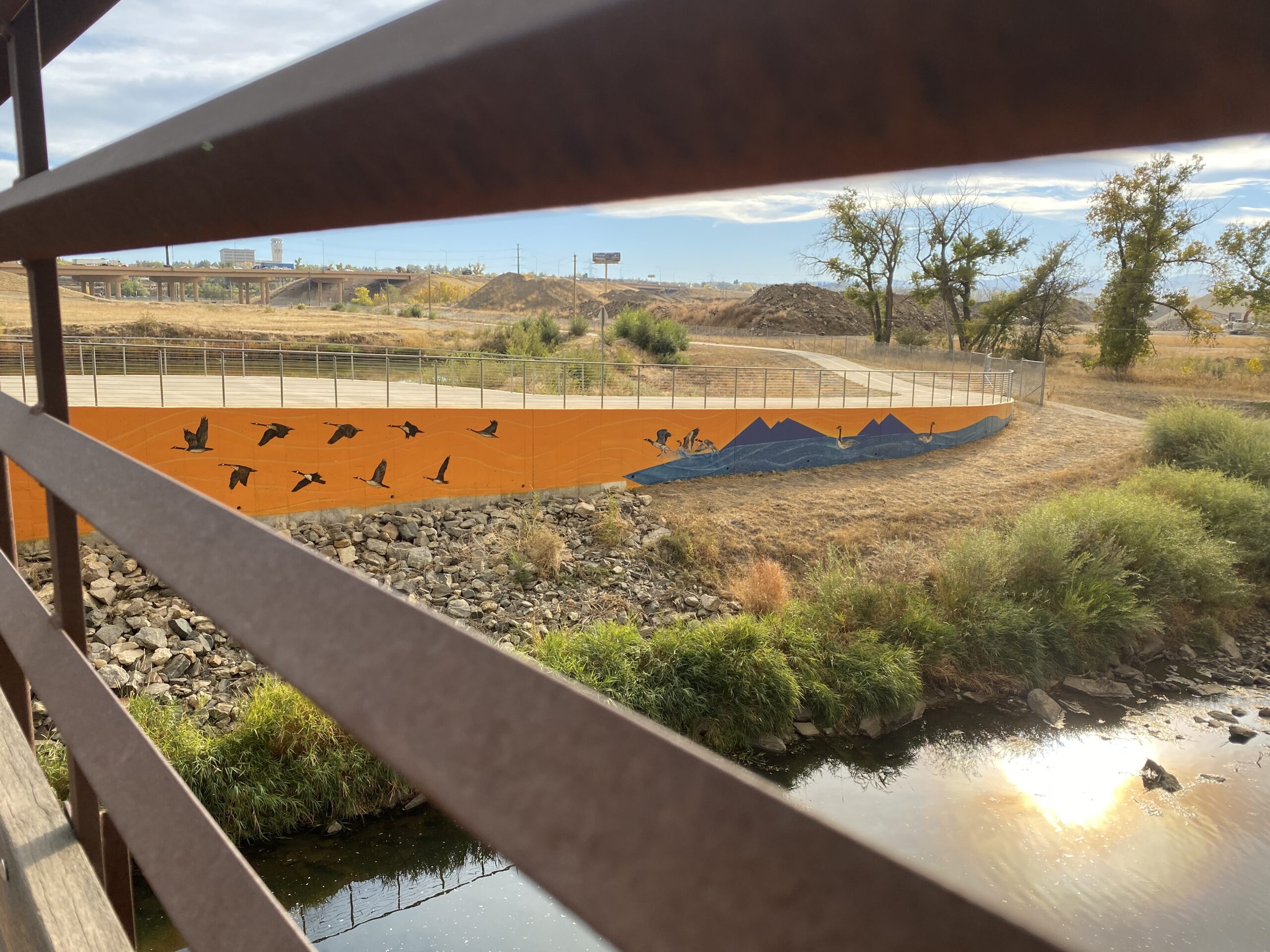 View from bridge showing a mural of flying birds and mountains on an orange wall along a dry, grassy landscape with a small river below.