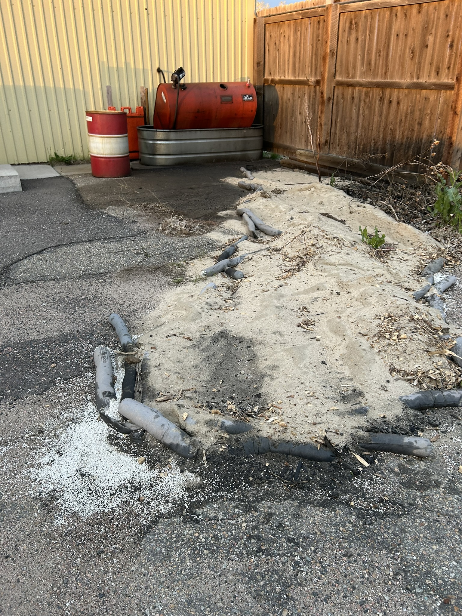 Sand pile with containment hoses near red fuel tanks and wooden fence