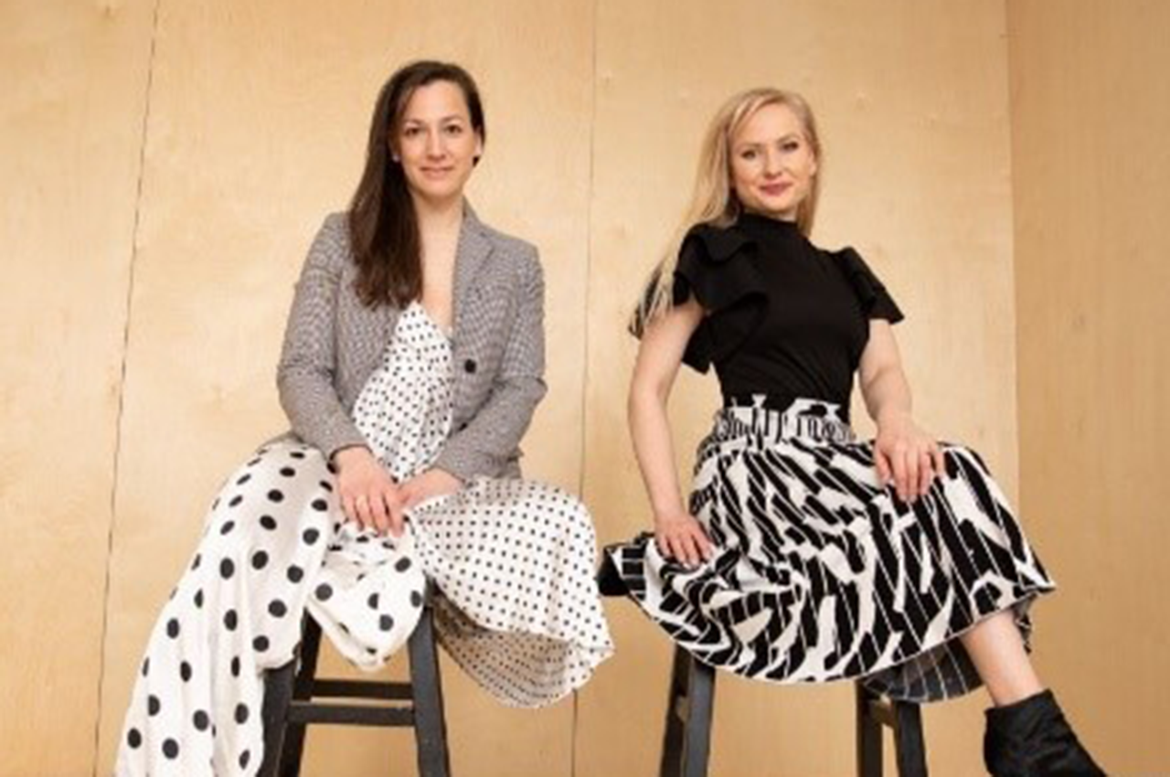 Two women sitting on stools, wearing polka dot and patterned skirts, smiling confidently.
