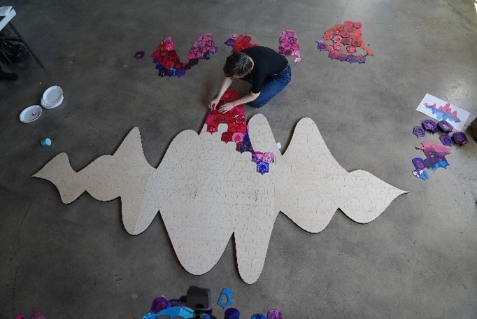 Mollie arranging colorful fabric pieces on large wooden wave shape in studio.