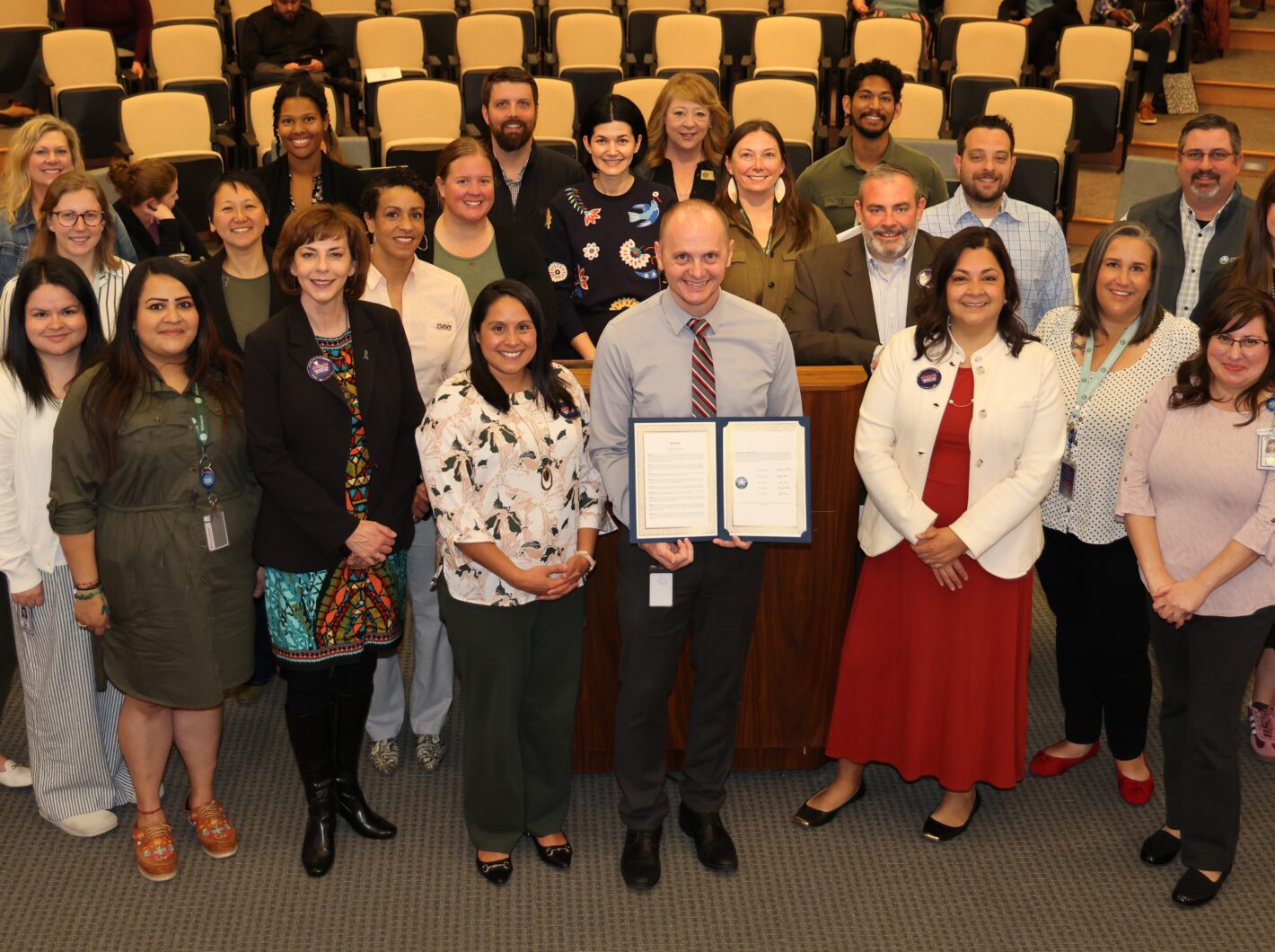 Group of colleagues smiling, holding a framed document, celebrating National Public Health Week in a public hearing room