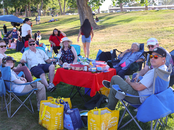 Group of people sitting in a park having a picnic with a red tablecloth.