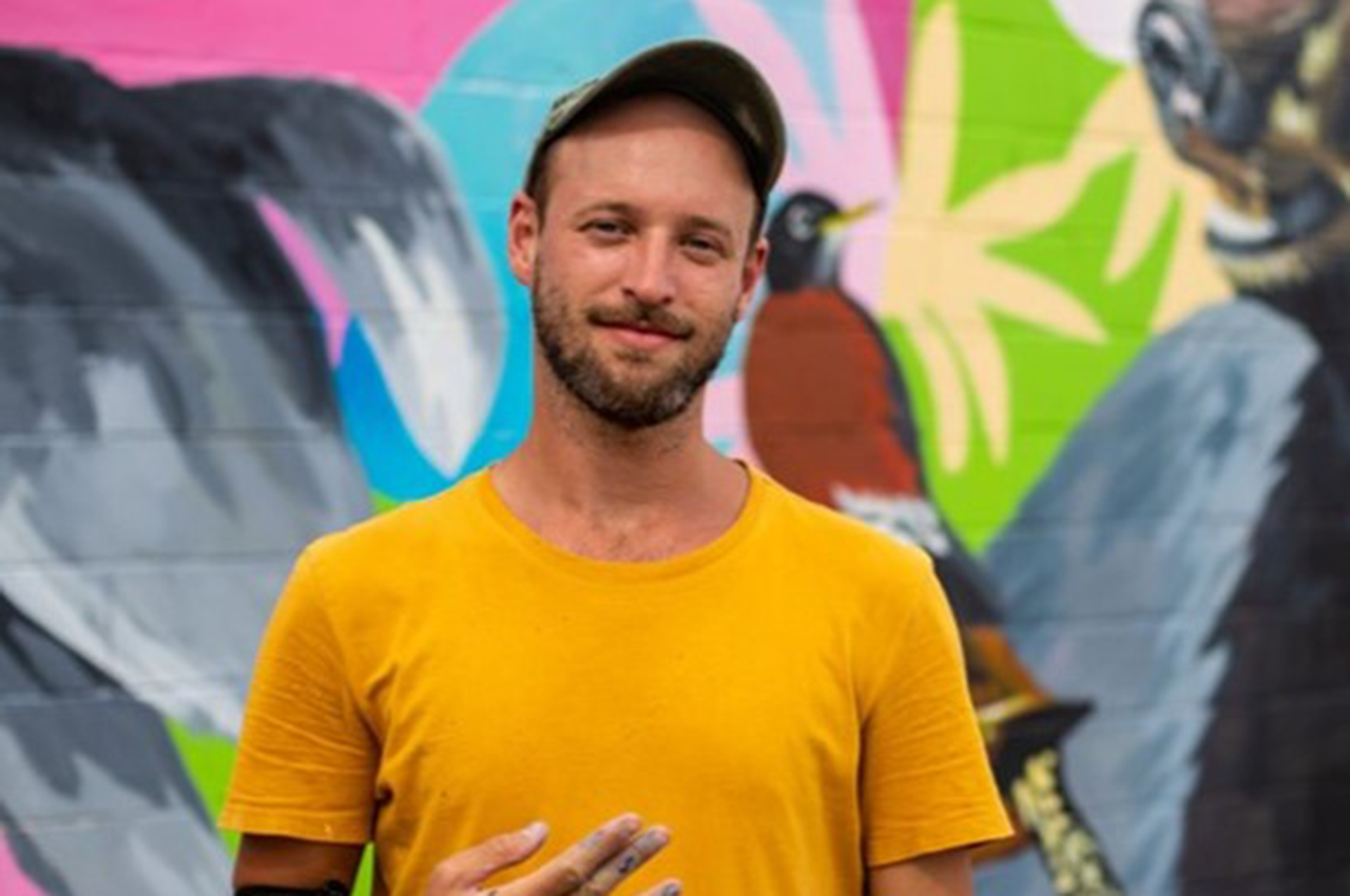 Primo in a yellow shirt standing before a colorful mural with a bird design