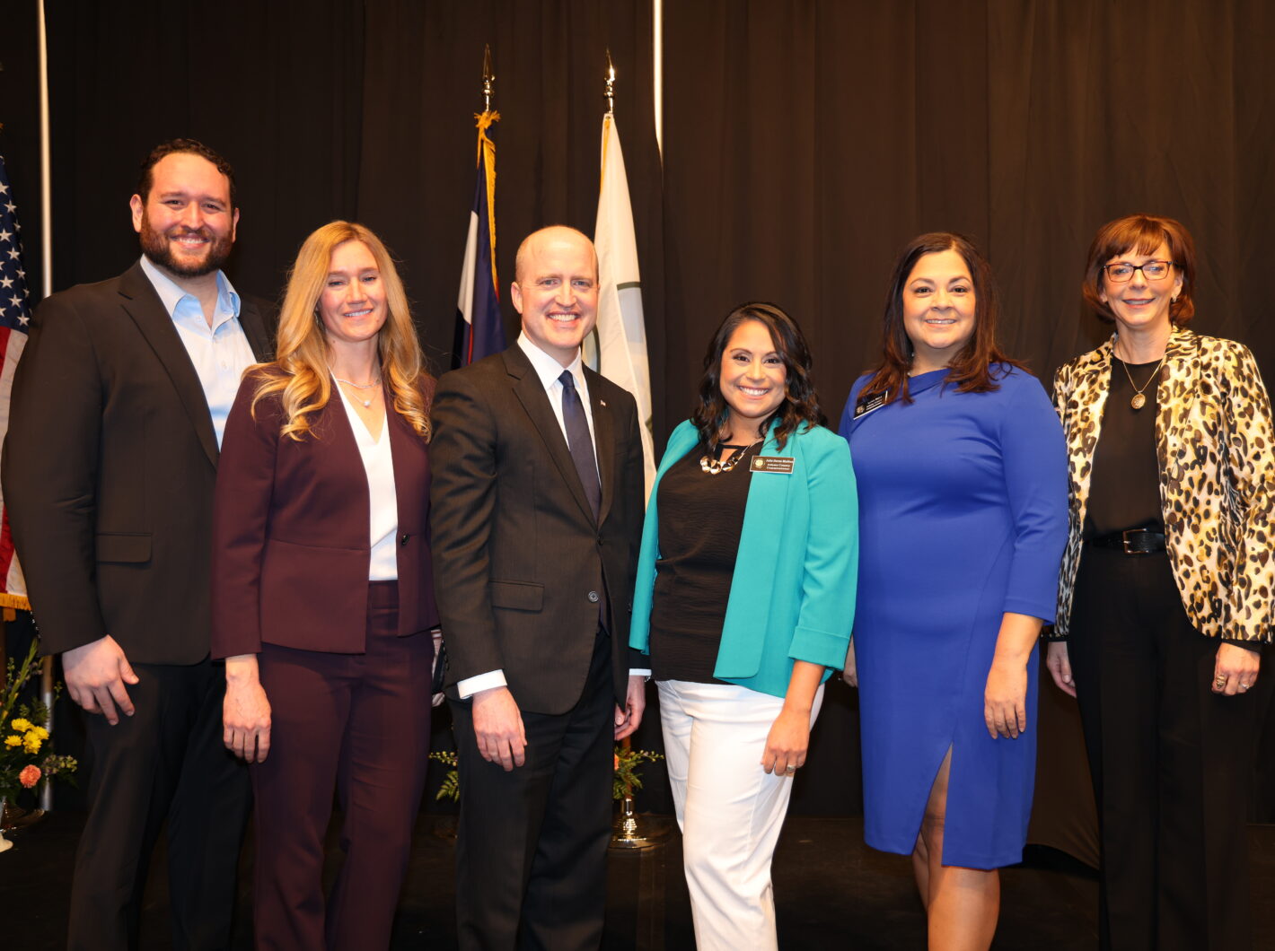 Six newly elected Adams County officials smiling, standing together with flags in the background, dressed in formal attire