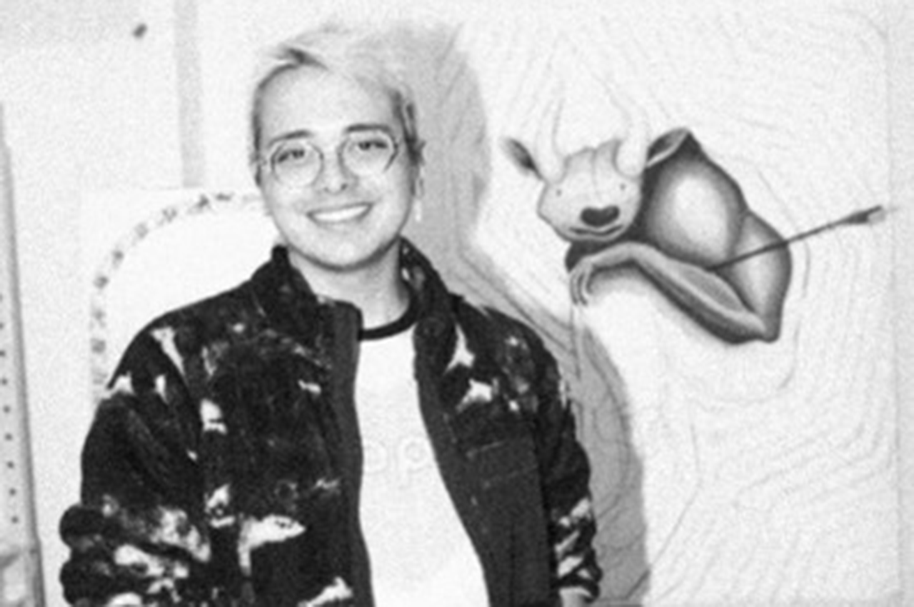 Vargass.png: Smiling person wearing glasses and a patterned jacket stands beside animal artwork.