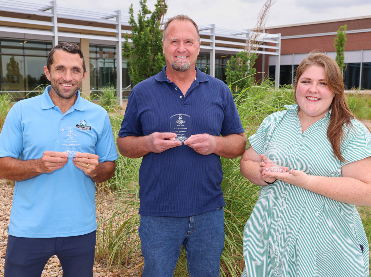 Three people holding awards, smiling outdoors in a garden setting.