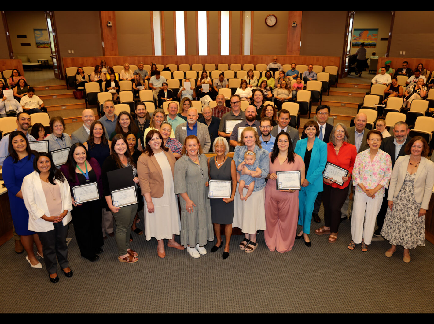 Group of people in a public hearing room, some holding certificates, smiling at the camera
