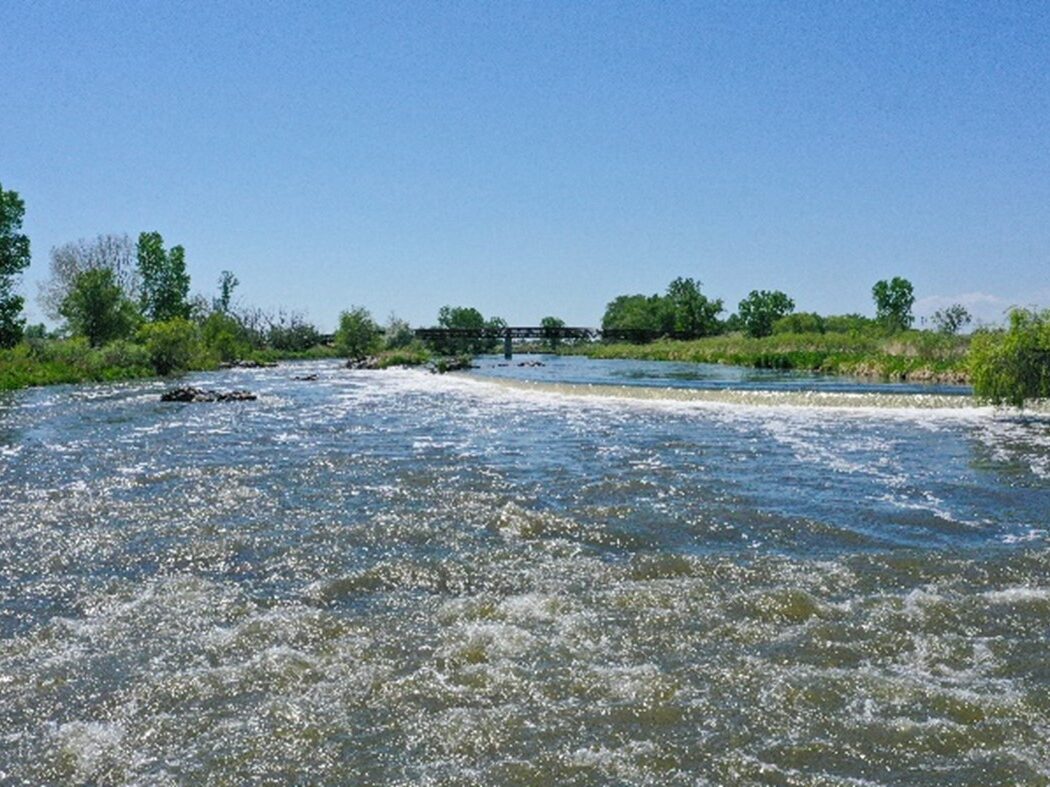 Flowing river with trees and a distant bridge under clear sky